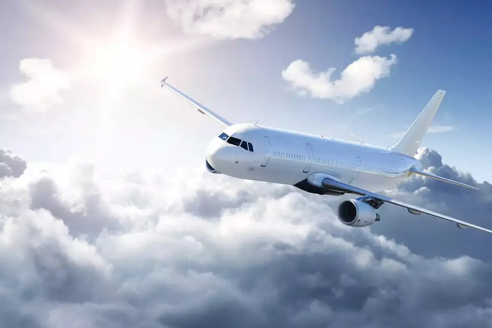 Un avión de pasajeros blanco volando sobre nubes suaves en un cielo brillante, con la luz del sol brillando desde la parte superior izquierda.