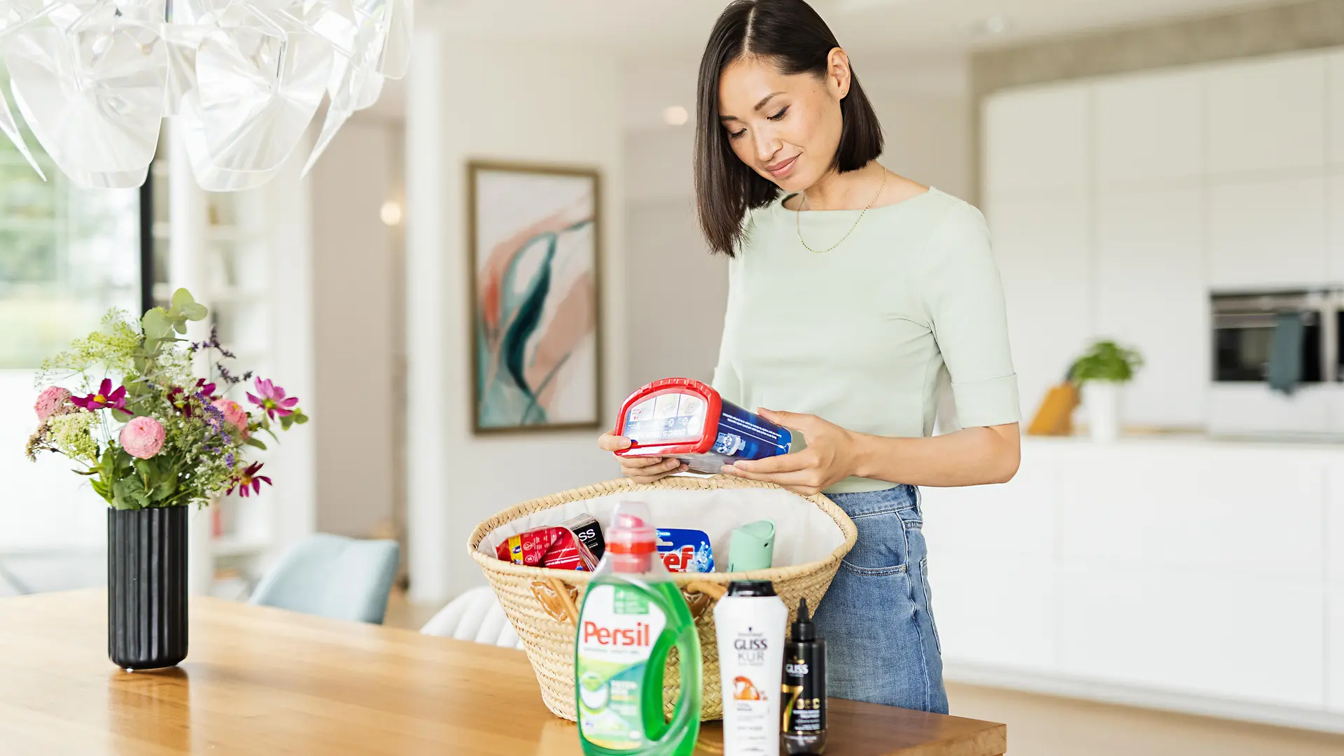 Mujer de pie con una cesta sobre una mesa que está llena de productos Henkel como: Persil, Gliss, Bref, Somat, taft y más.