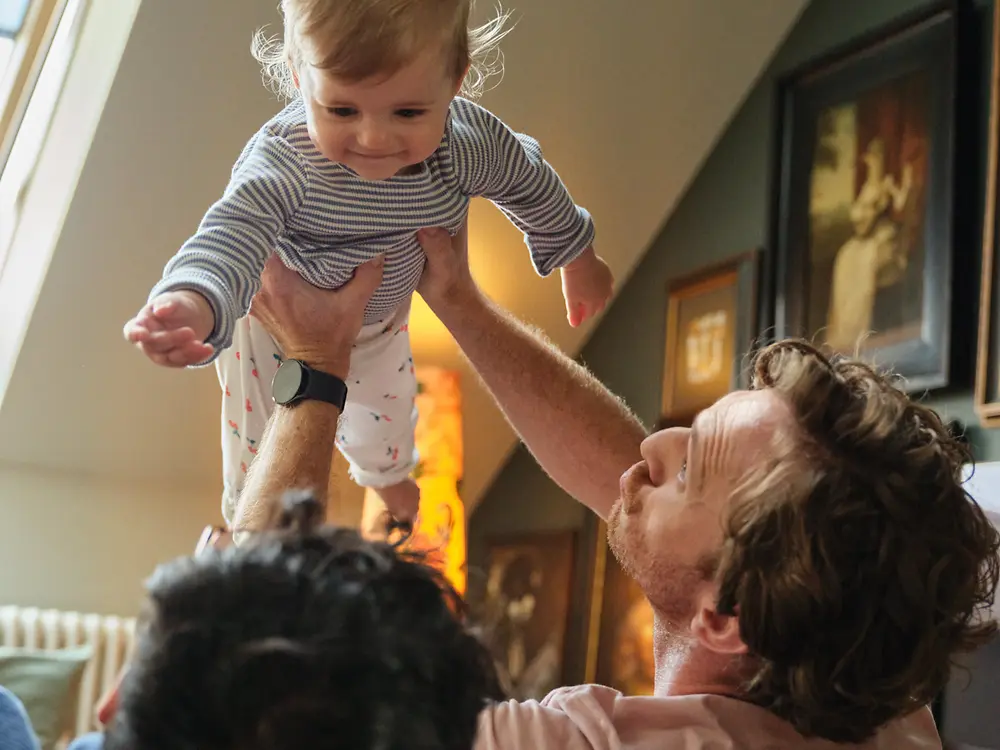 Dos padres sostienen a su bebé y sonríen en casa, lo que simboliza la inclusión y la política de licencia parental de Henkel, favorable para la familia.