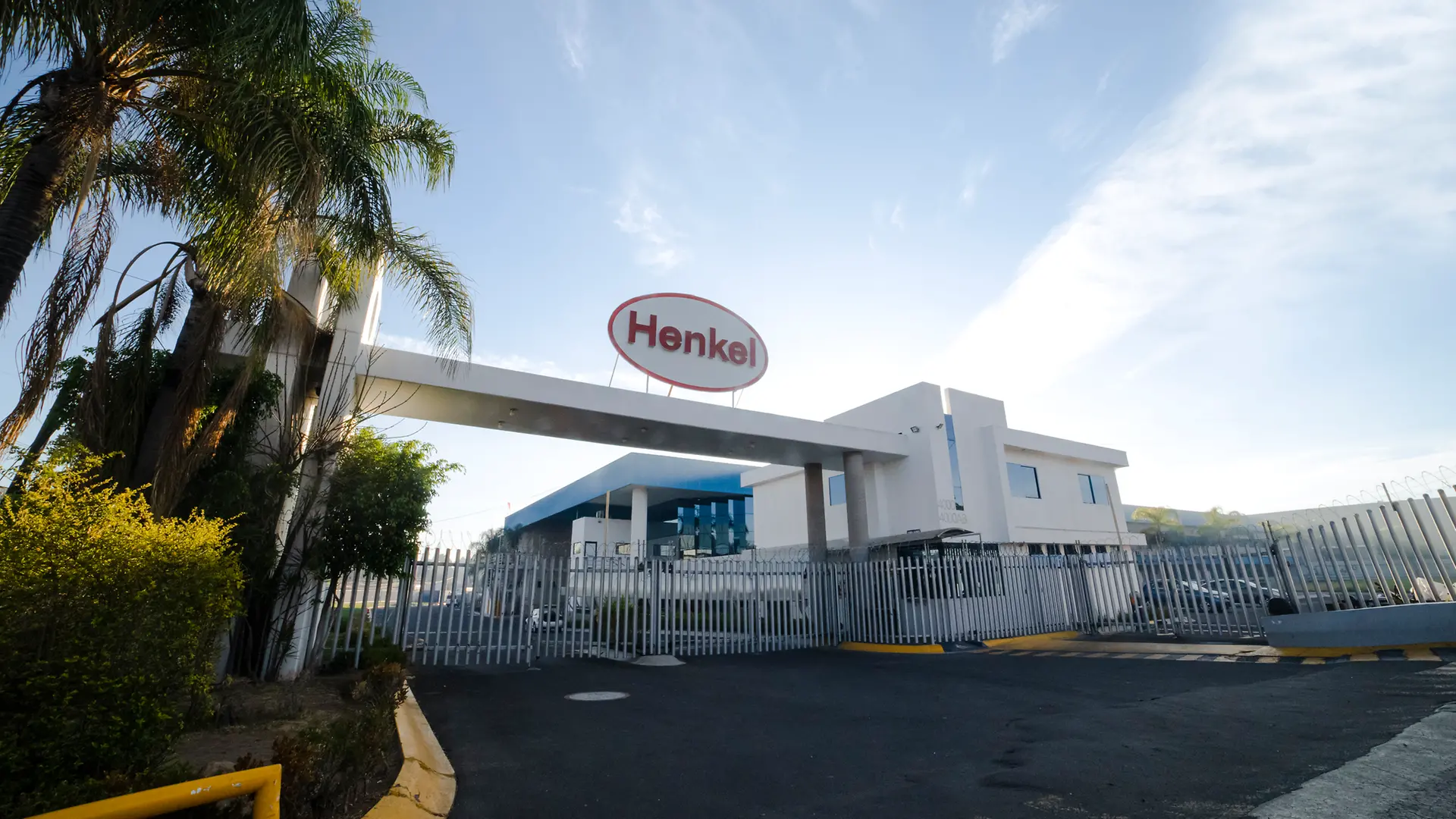 Entrada principal de planta Tesistán con estructura blanca y portón metálico, rodeada de palmeras y vegetación.