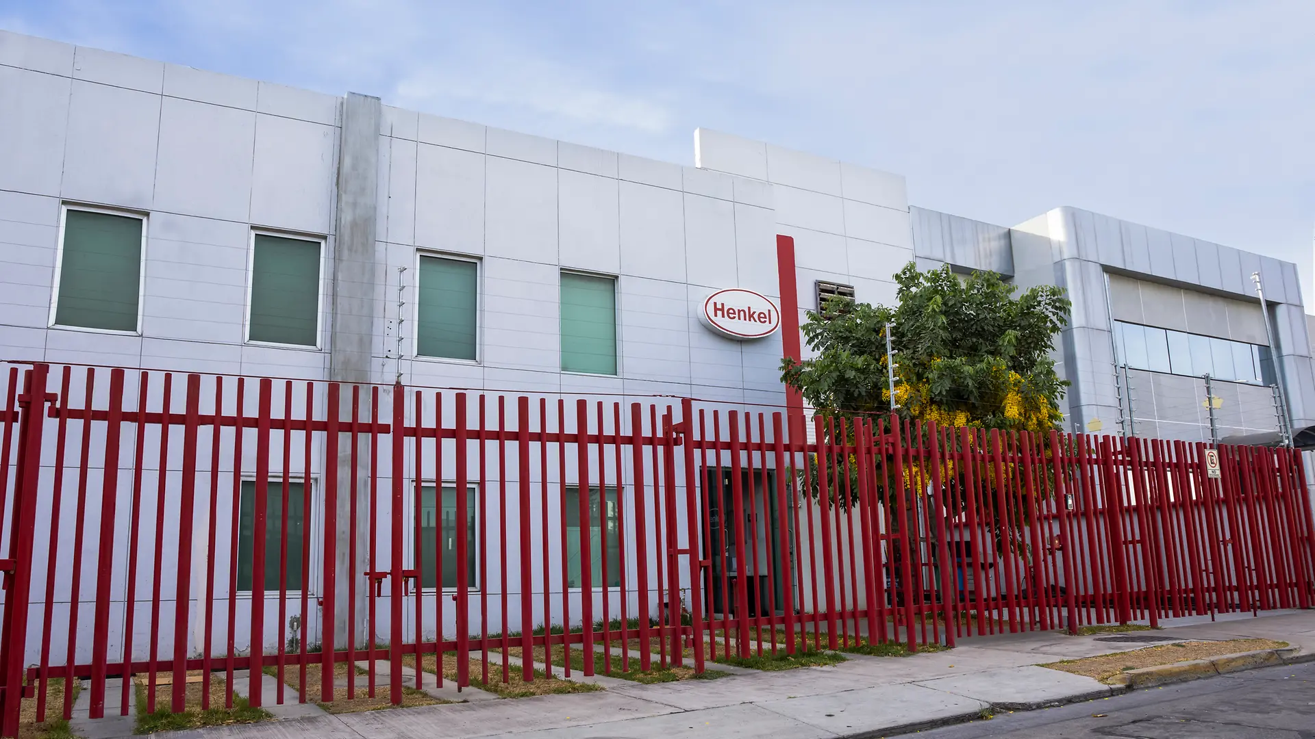 Edificio planta Natco de dos niveles con fachada blanca y reja metálica roja al frente. Árbol y acera visibles en la parte inferior.