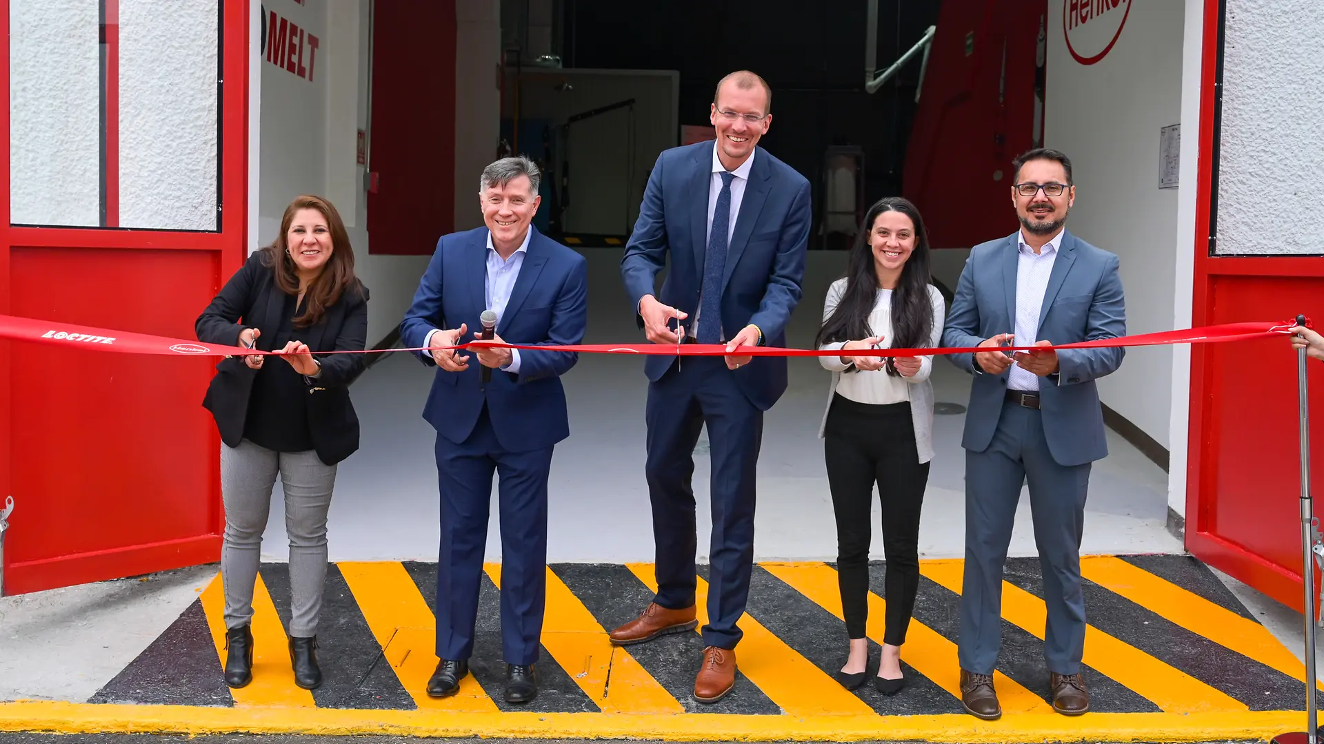 Ceremonia de inauguración con cinco personas cortando una cinta roja frente a una entrada industrial con puertas rojas y el logotipo de Henkel en la pared.