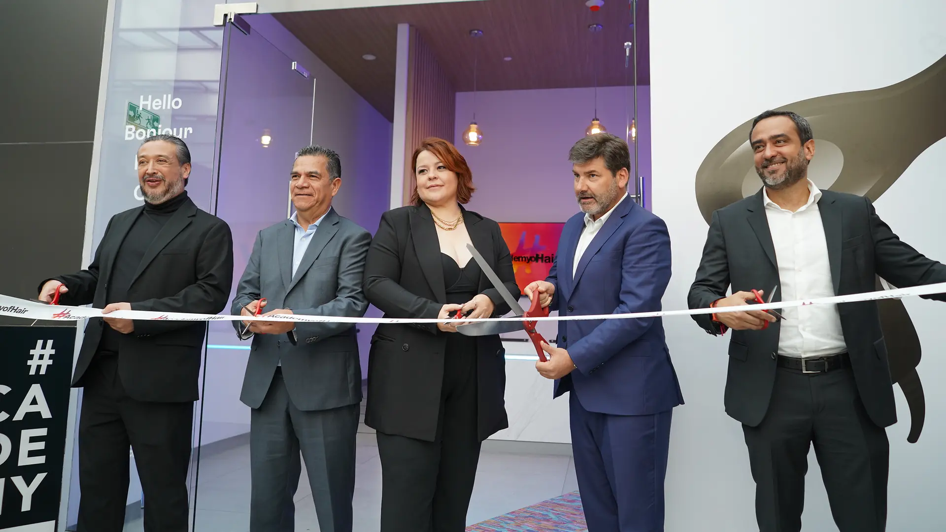 Ceremonia de inauguración con cinco personas cortando una cinta blanca frente a una entrada acristalada. En el fondo se observan lámparas colgantes y decoración interior con tonos cálidos.