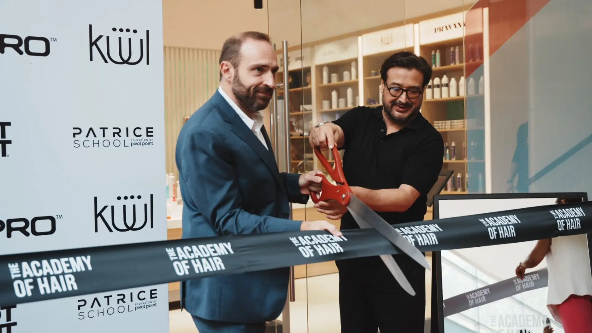 Dos personas cortando una cinta negra con el texto “THE ACADEMY OF HAIR” frente a una entrada de tienda con estanterías de productos visibles. A la izquierda hay un panel con logotipos de Patrice School, KÜÜL y otras marcas.