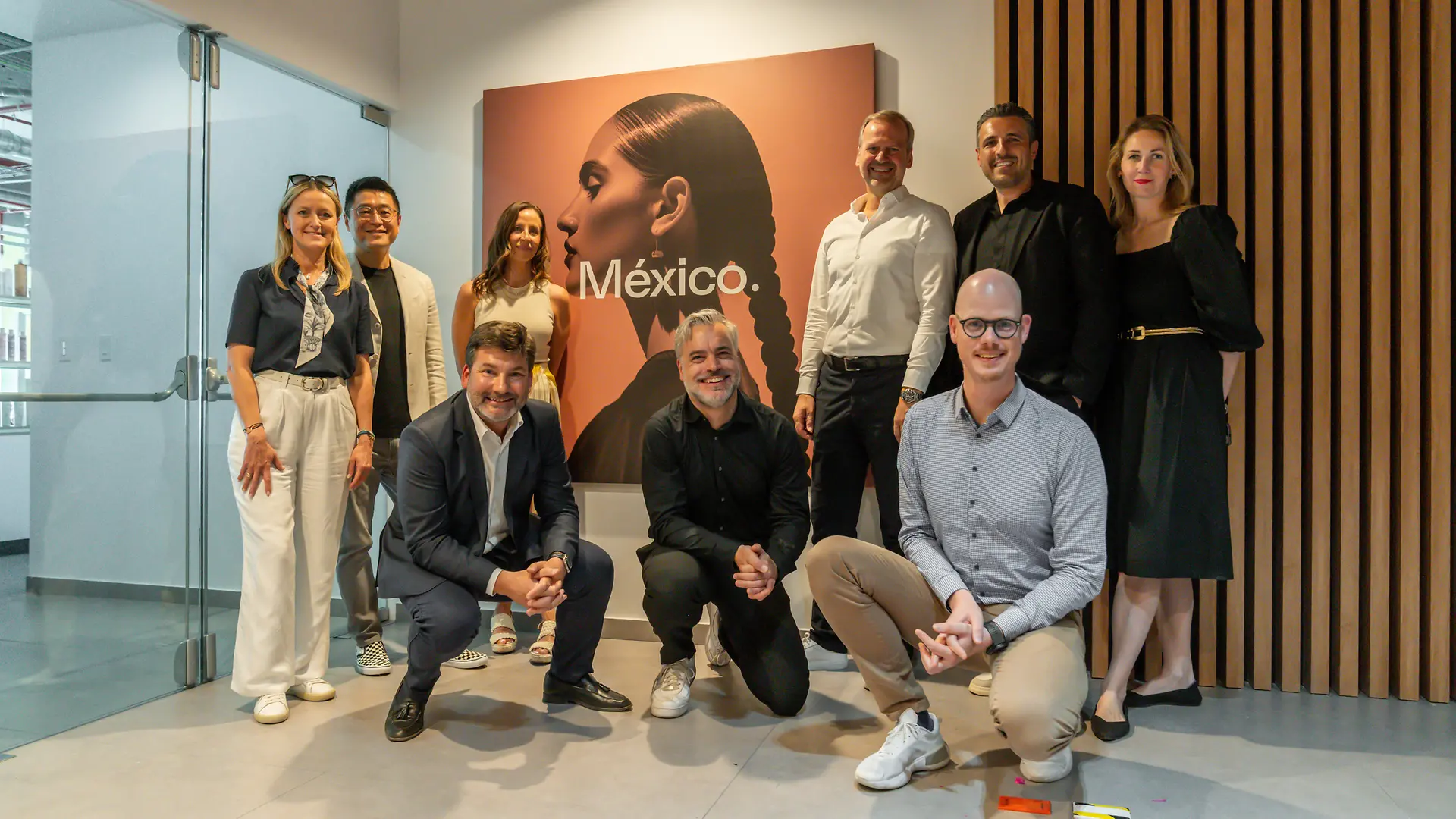 Grupo de personas posando en un espacio interior moderno frente a un cuadro con la palabra “México” y una imagen artística de una persona con trenza. Fondo con pared blanca y paneles de madera verticales.