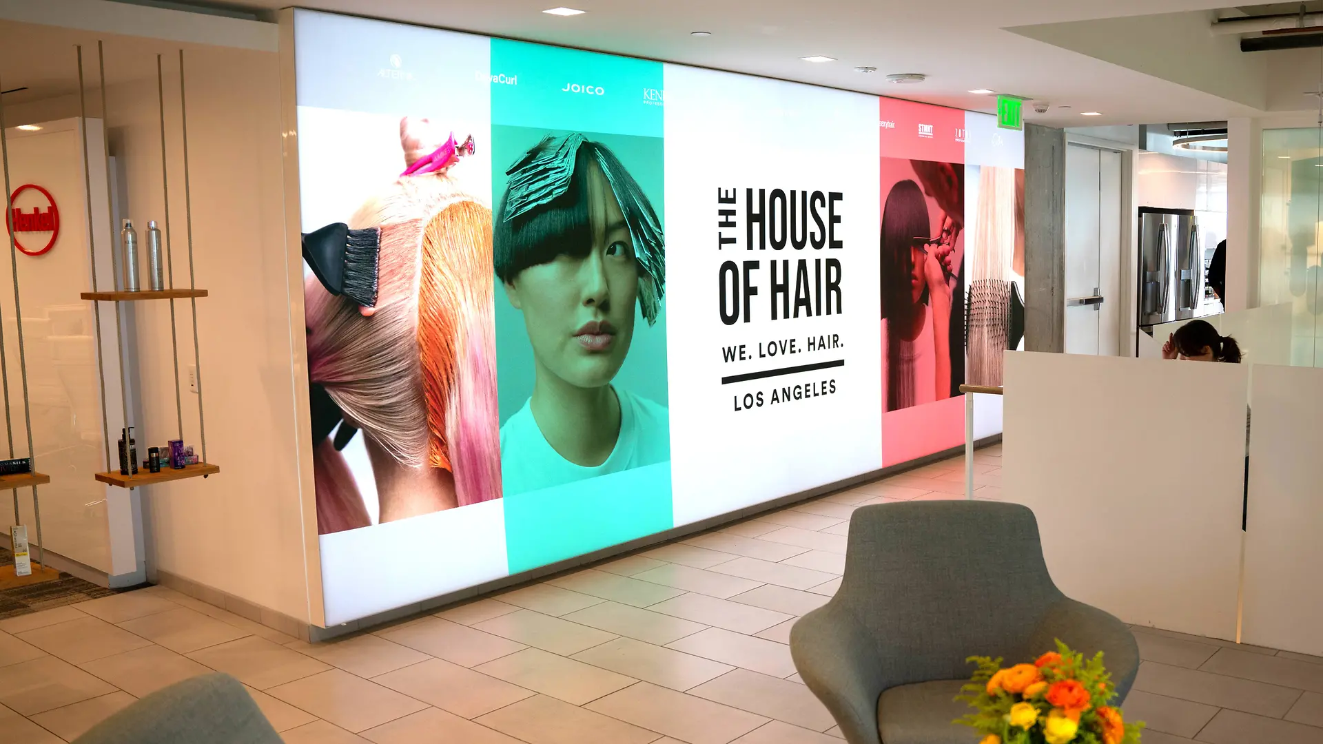 Vestíbulo moderno con una llamativa pared de exhibición que presenta grandes paneles con gráficos coloridos inspirados en el cabello y el texto: “THE HOUSE OF HAIR – WE. LOVE. HAIR. – LOS ANGELES”. El espacio incluye sillones lounge en gris claro, una pequeña mesa con un vibrante arreglo floral y un diseño limpio y minimalista con piso de baldosas y divisiones blancas.
