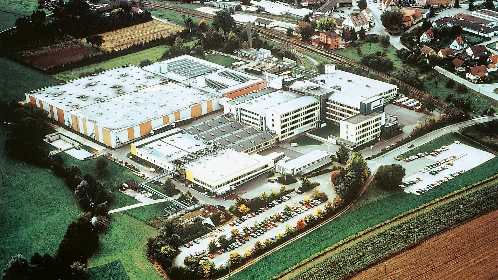 Vista aérea de la planta de Schwarzkopf en Wassertrüdingen en 1995, donde se muestran varios edificios y naves de producción, así como un gran estacionamiento lleno de autos.