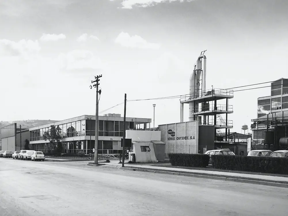 ista frontal en blanco y negro de una planta industrial con edificios de oficinas, estructuras metálicas y automóviles estacionados, con un letrero que indica “Henkel Onyx Mex, S.A.” en la entrada.