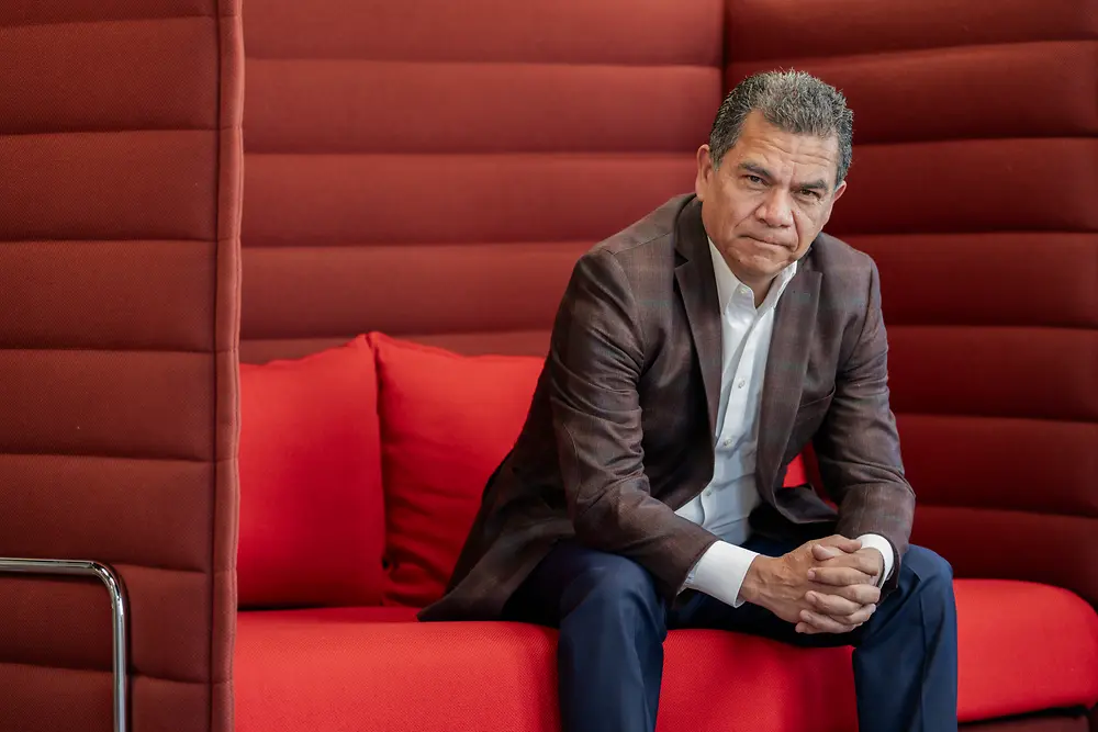 Persona sentada en un sillón acolchado de color rojo, vistiendo saco oscuro y pantalones formales, en un área interior de diseño corporativo.