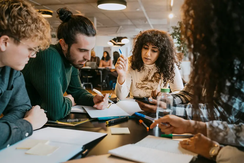 Un grupo de personas sentadas alrededor de una mesa en un espacio de trabajo interior moderno, colaborando con documentos, cuadernos y dispositivos digitales. Sobre la mesa hay varios bolígrafos, papeles y notas adhesivas. 