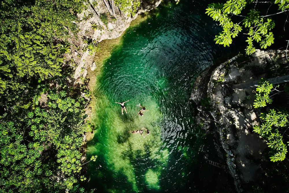 Vista aérea de una pequeña piscina natural rodeada de densa vegetación verde, con tres personas flotando en el agua clara de color esmeralda. 