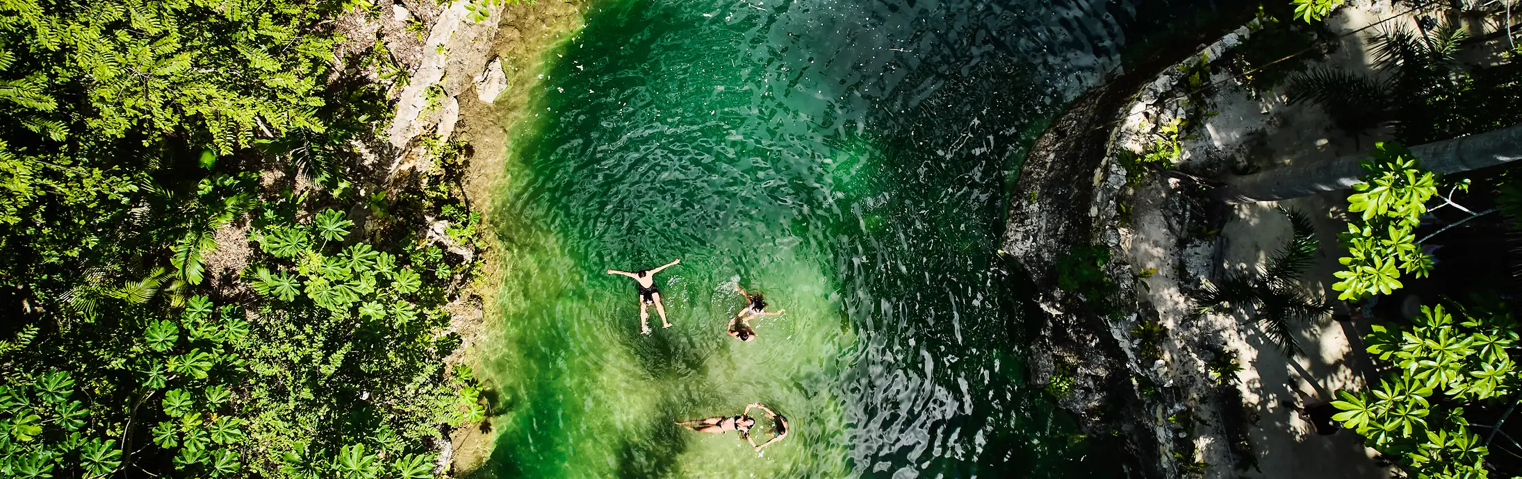 Vista aérea de una pequeña piscina natural rodeada de densa vegetación verde, con tres personas flotando en el agua clara de color esmeralda. 