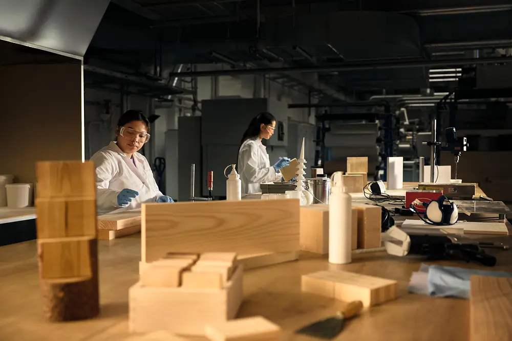 Entorno de taller con personas que llevan batas de laboratorio y guantes de protección, trabajando en una gran mesa de madera cubierta con muestras de madera, herramientas y botellas. 