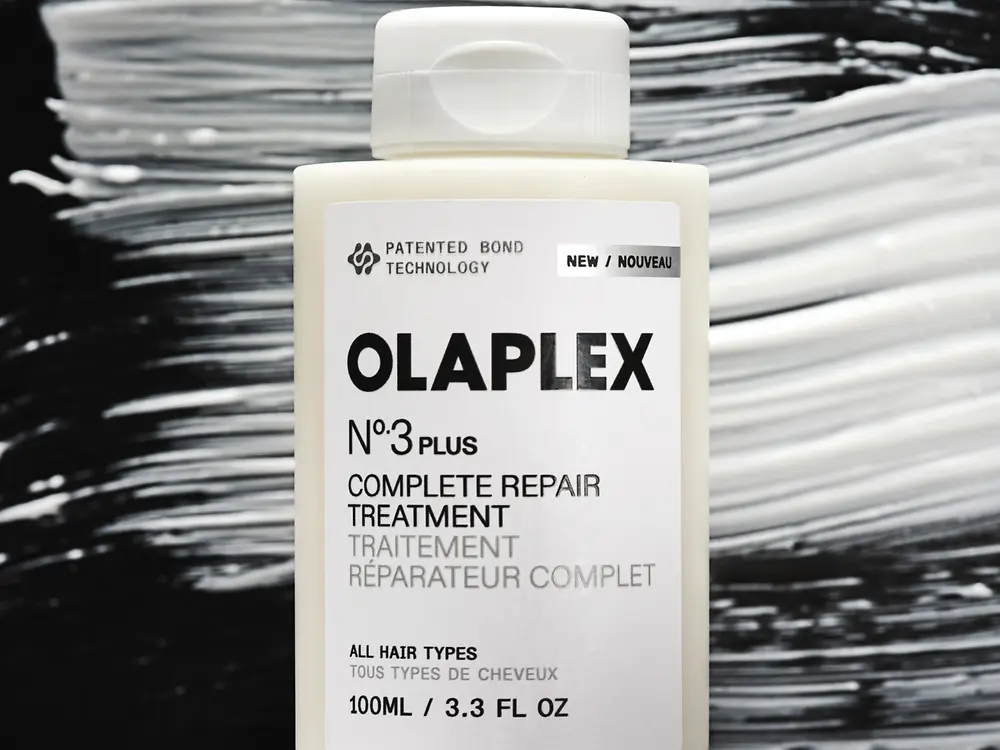 Fotografía de producto que muestra un frasco blanco de “OLAPLEX N°3 Plus Complete Repair Treatment” con tapa abatible. El fondo presenta trazos en tonos blanco, gris y negro con apariencia de pinceladas horizontales.