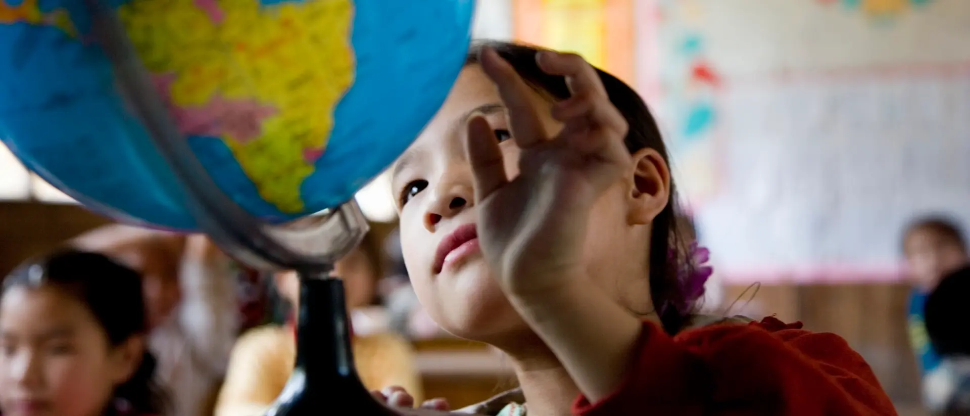 Un niño en un aula extiende la mano hacia un gran globo terráqueo colocado sobre un escritorio, con mapas coloridos y carteles visibles en las paredes al fondo. 