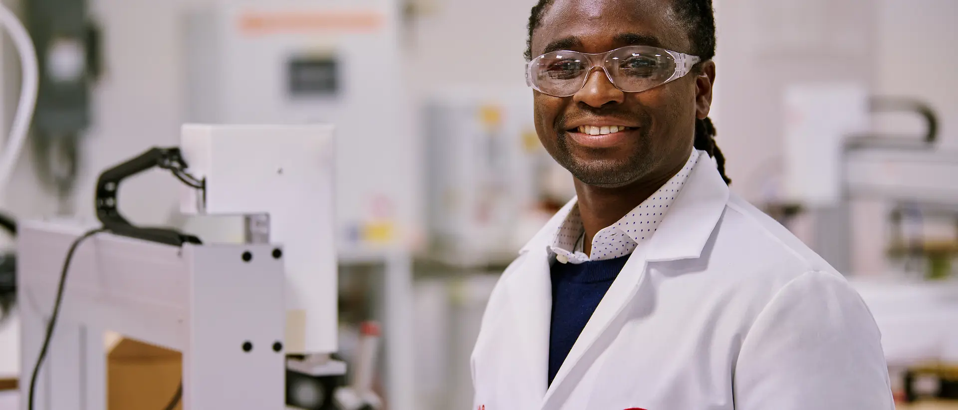 Una persona con una bata blanca de laboratorio de Henkel, de pie en un entorno de laboratorio rodeado de equipos y maquinaria. 