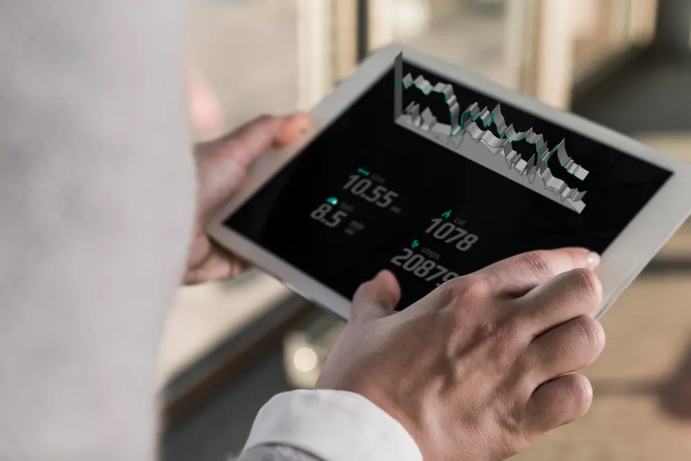 Una persona sostiene una tableta que muestra gráficos digitales y datos numéricos, vista por encima del hombro en un entorno interior luminoso. 