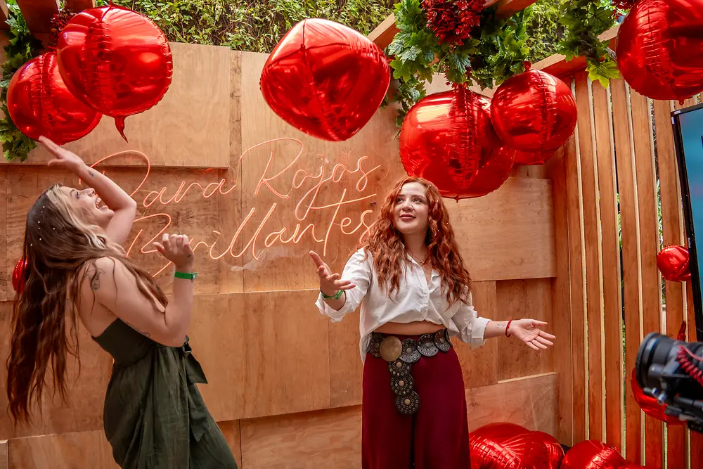 Dos personas participan en una dinámica frente a un fondo de madera con letrero luminoso. Alrededor hay globos rojos en forma de corazón.