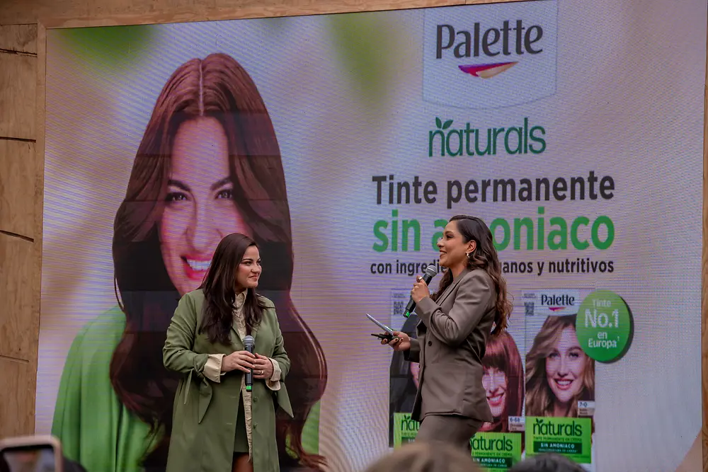 Dos personas en un escenario sostienen micrófonos frente a una pantalla grande con la marca “Palette Naturals” y el texto “Tinte permanente sin amoniaco”. En el fondo aparecen imágenes de productos y retratos de modelos con distintos tonos de cabello.