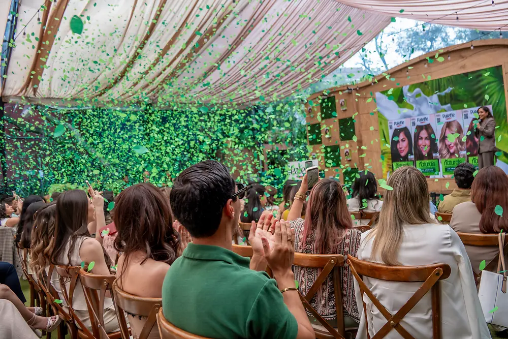 Audiencia sentada en sillas de madera durante un evento al aire libre, bajo una carpa decorada. Al frente hay un escenario con pantallas que muestran productos de la línea “Palette Naturals”.
