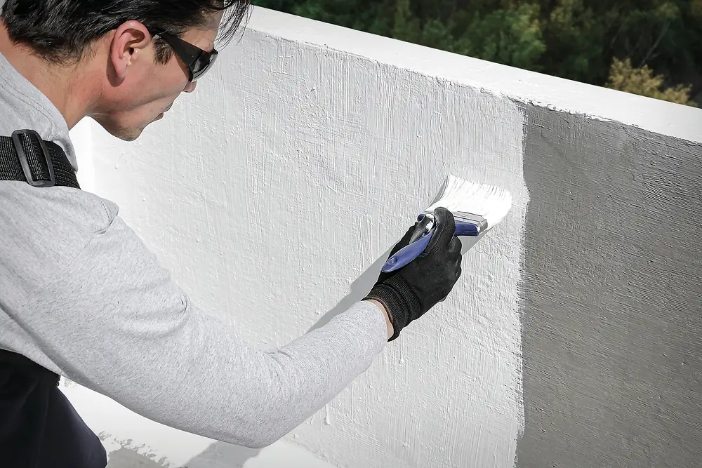 Primer plano de una persona utilizando una brocha para aplicar recubrimiento blanco Fester sobre un muro exterior. La brocha deja una capa visible y uniforme del material.