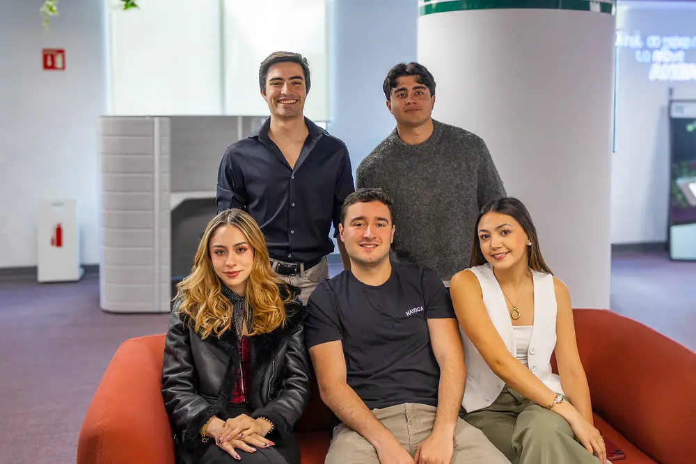 Grupo de cinco colegas reunidos alrededor de un sofá rojo en una moderna sala de descanso de la oficina, posando para una foto de equipo.