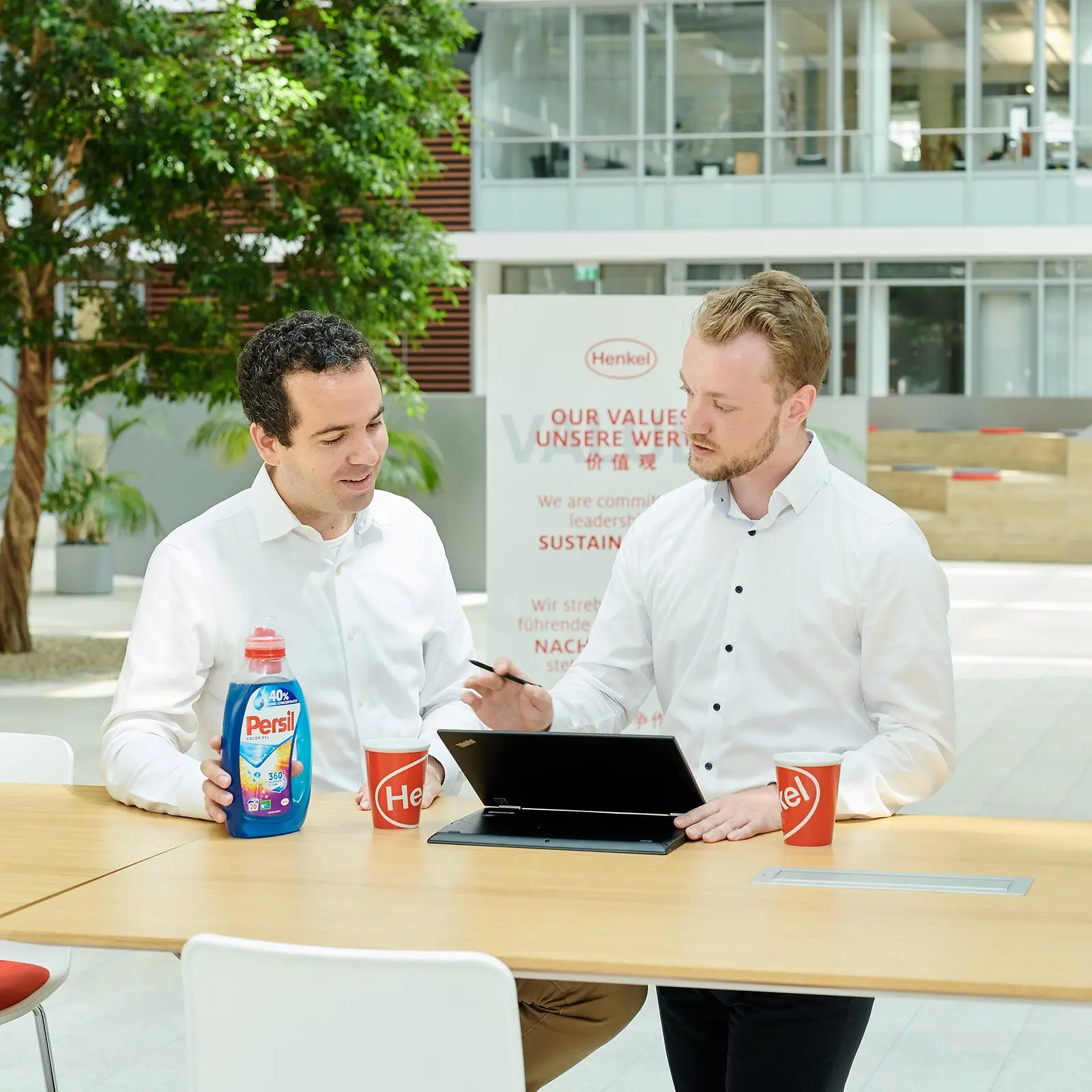Dos hombres de pie a lado de una mesa con una botella de Persil y una computadora portátil.